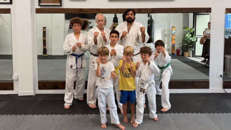 Instructor John and guest instructor Michael posing with K.I.C.K. students in karate uniforms with fists up.
